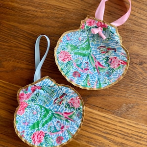 May include: Two seashell ornaments with a gold trim and a colorful nautical pattern. The ornaments have a blue, pink, and green design with a pink ribbon for hanging. The background is a wooden surface.