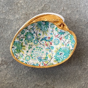 May include: A decorative clam shell with a gold-painted rim and a floral interior. The interior features a white background with teal, yellow, and blue floral and bird patterns. The shell is resting on a gray stone surface.