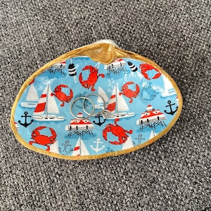 Puede incluir: Un plato para anillos en forma de concha de color azul y blanco con un borde dorado. El plato está decorado con cangrejos rojos, veleros, faros y anclas. Dos anillos de oro están colocados en el plato.