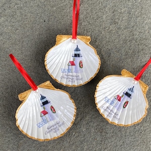 May include: Three seashell ornaments with a lighthouse design and the text "Nantucket Christmas Stroll 2025". Each ornament has a red ribbon for hanging and a gold-painted edge.