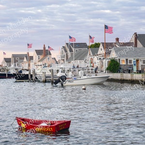 May include: A red rowboat with the words "Sunken Ship" and "Point Judith" on the side is docked in a harbor with several white boats and houses in the background. American flags are flying from the houses.