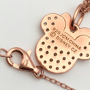 May include: A rose gold-toned necklace featuring a Minnie Mouse-shaped pendant with a grid of small circular cutouts. The back of the pendant is engraved with 925 JCM CHINA DISNEY CZ. A lobster claw clasp with 925 and JCM markings is attached.