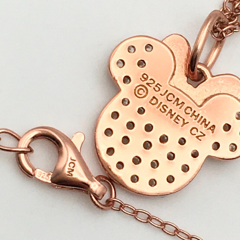 May include: A rose gold-toned necklace featuring a Minnie Mouse-shaped pendant with a grid of small circular cutouts. The back of the pendant is engraved with 925 JCM CHINA DISNEY CZ. A lobster claw clasp with 925 and JCM markings is attached.