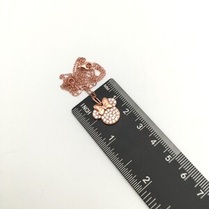 May include: A rose gold-toned necklace featuring a pendant shaped like a mouse head with a bow, encrusted with clear stones. The pendant measures approximately 0.5 inches in width, as shown against a black ruler with imperial markings.