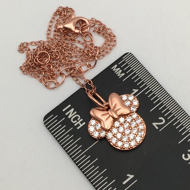 May include: A rose gold-toned necklace featuring a pendant shaped like a mouse head with a bow, encrusted with clear stones. The pendant is positioned on a black ruler showing a width of approximately 0.75 inches for the mouse head charm.