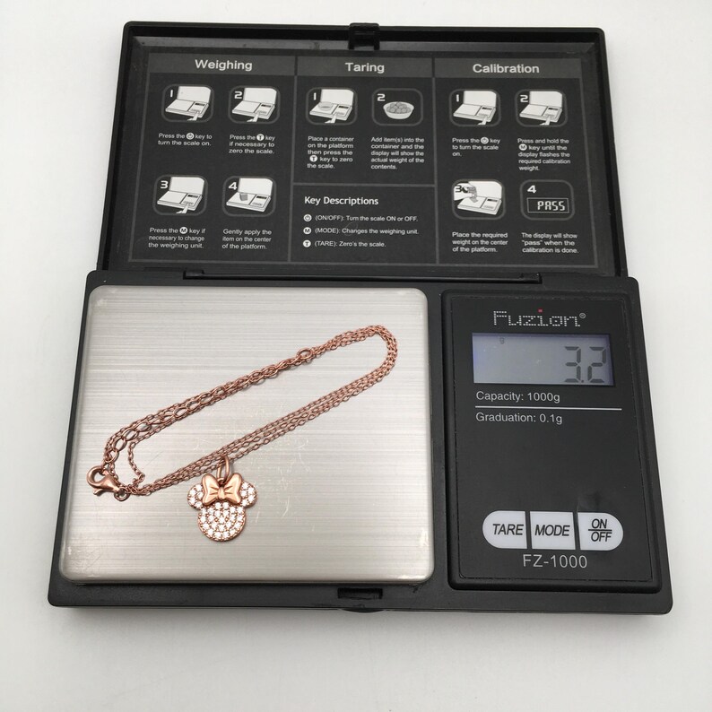 May include: A rose gold-toned chain bracelet with a Minnie Mouse-shaped charm featuring a bow and clear stones, resting on a digital scale that displays a weight of 3.2 grams. The scale has a black frame with white instructional text printed on it.