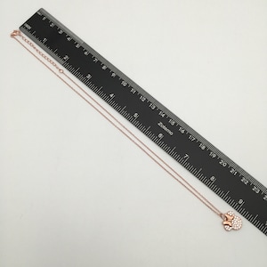 May include: A rose gold-toned necklace featuring a bow-shaped pendant encrusted with small clear stones. The necklace is positioned alongside a black ruler, showing a total length of approximately 9.5 inches from the clasp to the pendant.