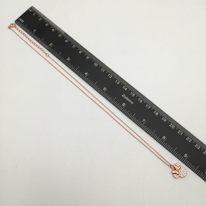 May include: A rose gold-toned necklace featuring a bow-shaped pendant encrusted with small clear stones. The necklace is positioned alongside a black ruler, showing a total length of approximately 9.5 inches from the clasp to the pendant.