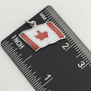 May include: A small, silver-toned charm in the shape of the Canadian flag. The flag features a white background with a red maple leaf in the center and red borders. The charm is approximately 1 inch in length and is shown next to a ruler.