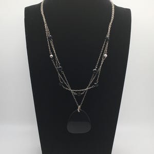 May include: A silver-toned necklace with three strands, featuring black beads and a large, black, teardrop-shaped pendant. The necklace is displayed on a black mannequin.