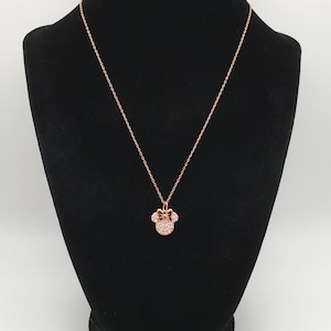 May include: A rose gold-toned necklace featuring a pendant shaped like a mouse head with a bow, encrusted with small clear stones. The pendant hangs from a delicate rose gold-toned chain, creating a simple and elegant piece of jewelry.