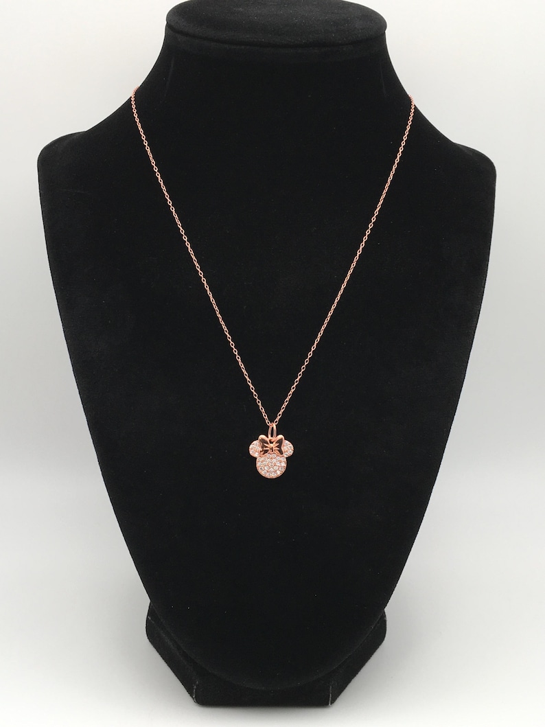 May include: A rose gold-toned necklace featuring a pendant shaped like a mouse head with a bow, encrusted with small clear stones. The pendant hangs from a delicate rose gold-toned chain, creating a simple and elegant piece of jewelry.