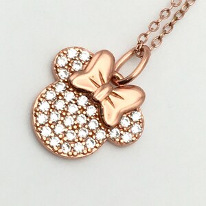 May include: A rose gold-toned necklace featuring a pendant shaped like a mouse head with a bow. The pendant is encrusted with numerous small, clear, sparkling stones covering the circular head portion, while the bow remains smooth metal.