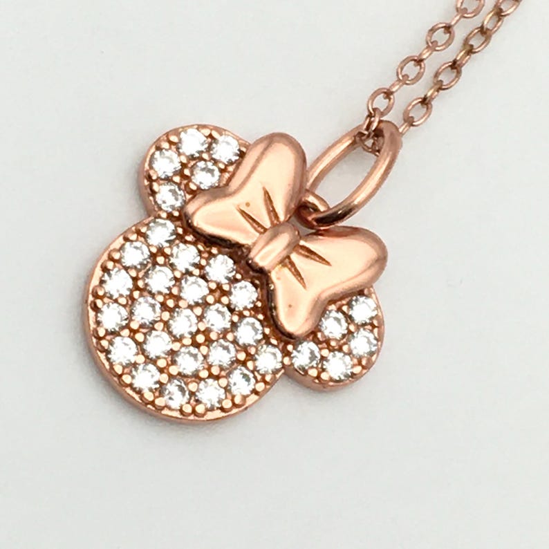 May include: A rose gold-toned necklace featuring a pendant shaped like a mouse head with a bow. The pendant is encrusted with numerous small, clear, sparkling stones covering the circular head portion, while the bow remains smooth metal.