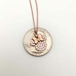 May include: A rose gold-toned necklace featuring a pendant shaped like a mouse head with a bow, encrusted with clear stones. The pendant is positioned on top of a United States quarter coin for scale, showing the relative size of the jewelry piece.