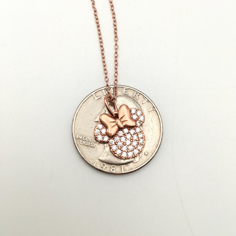 May include: A rose gold-toned necklace featuring a pendant shaped like a mouse head with a bow, encrusted with clear stones. The pendant is positioned on top of a United States quarter coin for scale, showing the relative size of the jewelry piece.