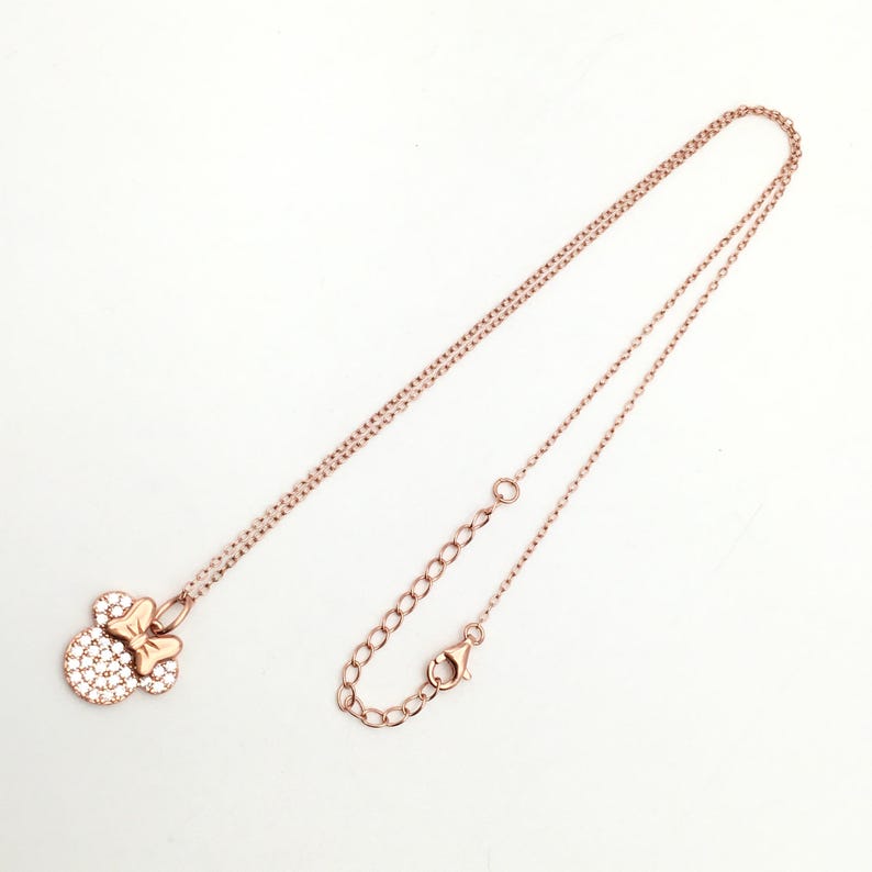 May include: A rose gold-toned necklace featuring a pendant shaped like a mouse head with a bow, embellished with small clear stones. The chain is thin and delicate, including an adjustable section with a lobster claw clasp at the end.