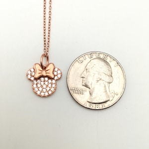 May include: A rose gold-toned necklace featuring a pendant shaped like a mouse head with a bow, encrusted with clear stones. The pendant is positioned next to a United States quarter coin for scale, showing the relative size of the jewelry piece.