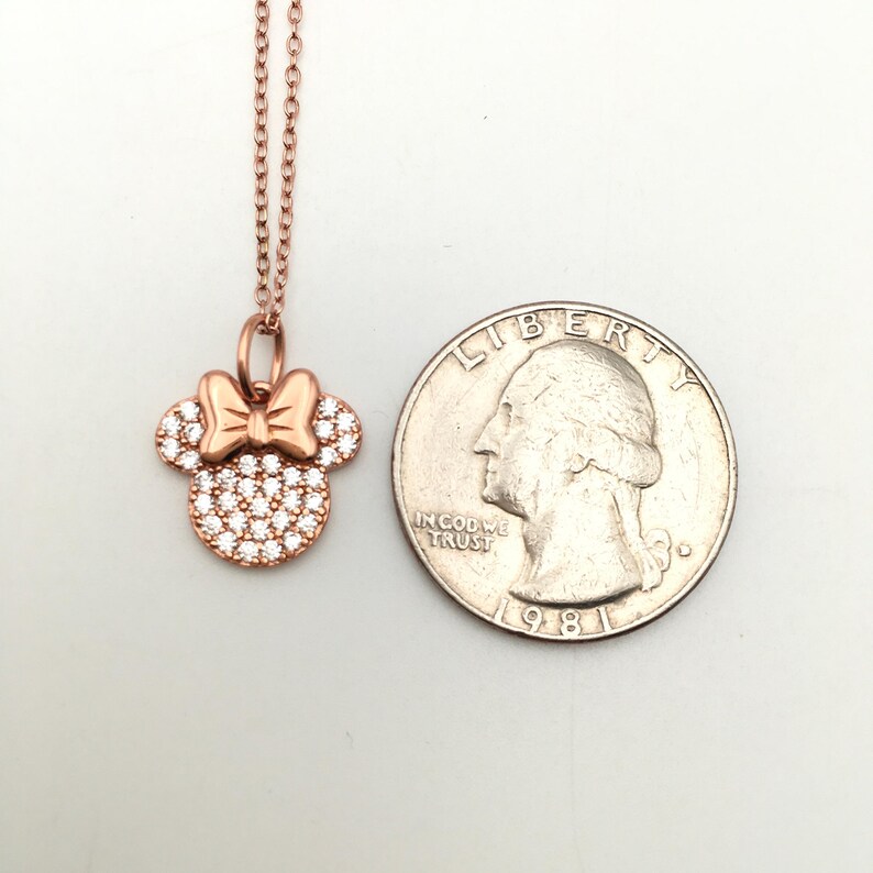 May include: A rose gold-toned necklace featuring a pendant shaped like a mouse head with a bow, encrusted with clear stones. The pendant is positioned next to a United States quarter coin for scale, showing the relative size of the jewelry piece.