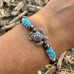 Op de afbeelding: Zilveren armband met een schildpadbedel, blauwe en zwarte geweven details. De armband heeft een gedetailleerde schildpadhanger en donkergrijze kralen. De blauwe geweven secties voegen een vleugje kleur toe.