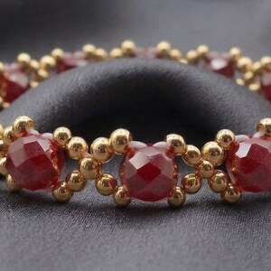 May include: A beaded bracelet featuring faceted, deep red gemstones and small gold-colored beads. The bracelet is arranged in a repeating pattern and rests on a dark gray, textured surface. The jewelry is designed to be worn as an accessory.