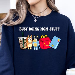 May include: Navy blue sweatshirt with the text "Busy Doing Mom Stuff" in white. The sweatshirt features a cartoon image of a dog holding a cup of coffee, a dog holding a blue toy, and a dog holding a red and yellow fast food container.