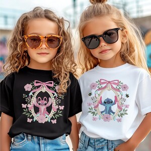 Pu&ograve; includere: Due bambine con occhiali da sole e magliette. Una maglietta &egrave; nera, l'altra bianca. Entrambe le magliette presentano un personaggio dei cartoni animati in una cornice floreale con un fiocco rosa. La maglietta nera ha un personaggio rosa e la maglietta bianca ha un personaggio blu.