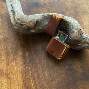 May include: A brown leather lighter case with a stitched design. The case is open and a silver lighter is visible inside.