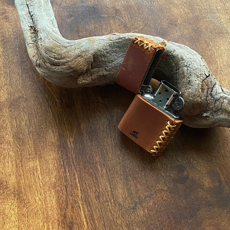 May include: A brown leather lighter case with a stitched design. The case is open and a silver lighter is visible inside.