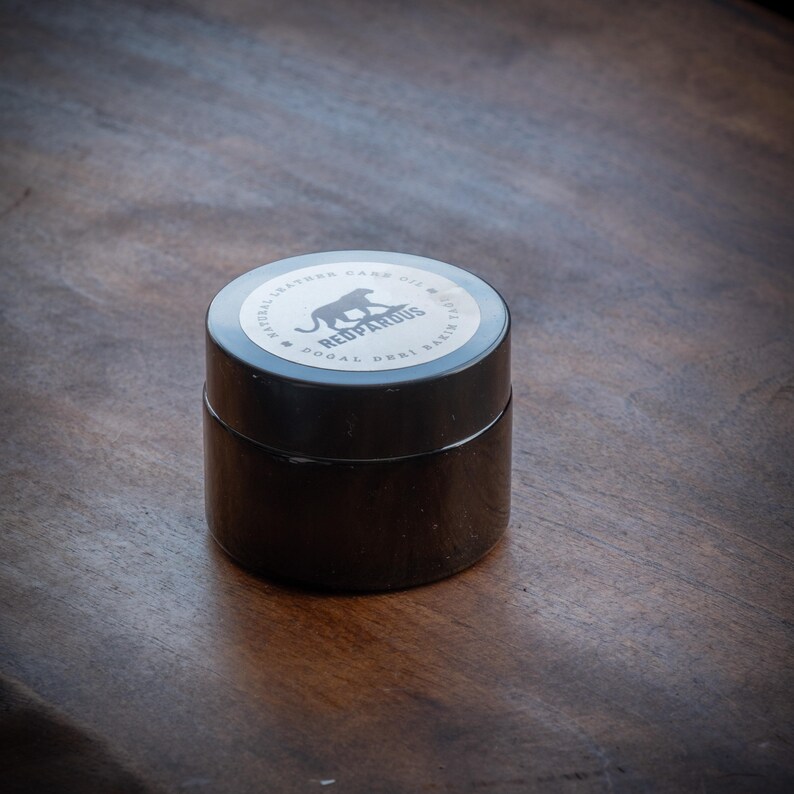May include: A black jar with a silver lid. The lid has a black panther silhouette and the text "NATURAL LEATHER CARE OIL - REDPANTROUS - DOOAL DERI BALSAM"