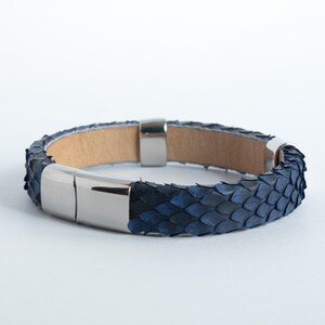 Puede incluir: Una pulsera de cuero azul y negro con un patrón de escamas. La pulsera tiene un cierre magnético de color plateado. El interior de la pulsera es de color marrón claro.