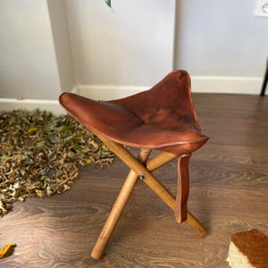 May include: A folding tripod stool featuring a triangular seat made of smooth, dark brown leather with visible stitching along the edges. The base consists of three cylindrical wooden legs that intersect at a central metal pivot point.