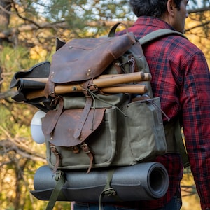 May include: A brown leather and green canvas backpack with a rolled-up yoga mat strapped to the bottom. The backpack has multiple pockets and straps, and is being worn by a person in a red and black plaid shirt.