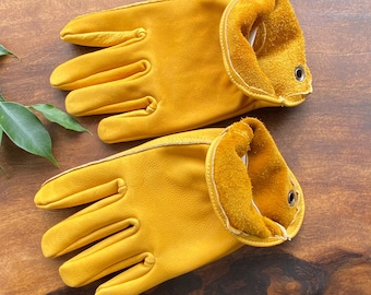 May include: A pair of yellow leather work gloves with a rolled cuff. The gloves have reinforced stitching and a suede-like interior cuff. The gloves are designed for manual labor and are shown on a wooden surface.