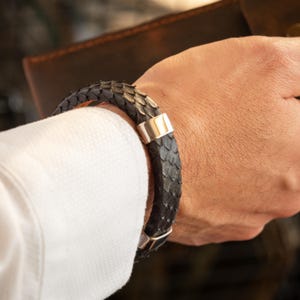 Può includere: Un bracciale in pelle di serpente grigio scuro con dettagli in metallo argentato. Il bracciale è indossato su un polso, in contrasto con una camicia bianca. La texture e il design del bracciale suggeriscono lusso e stile.