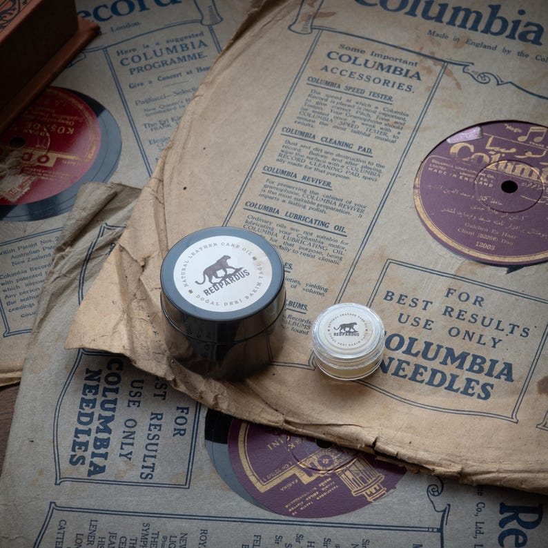 May include: Two small jars of record care products, one labeled "Redpardus Natural Leather Grease Oil" and the other with a red panther logo. The jars are sitting on top of old vinyl record sleeves with text that reads "Columbia" and "For Best Results Use Only Columbia Needles".