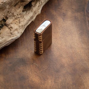 May include: A brown leather case with a silver metal insert and decorative stitching. The case is sitting on a wooden surface.