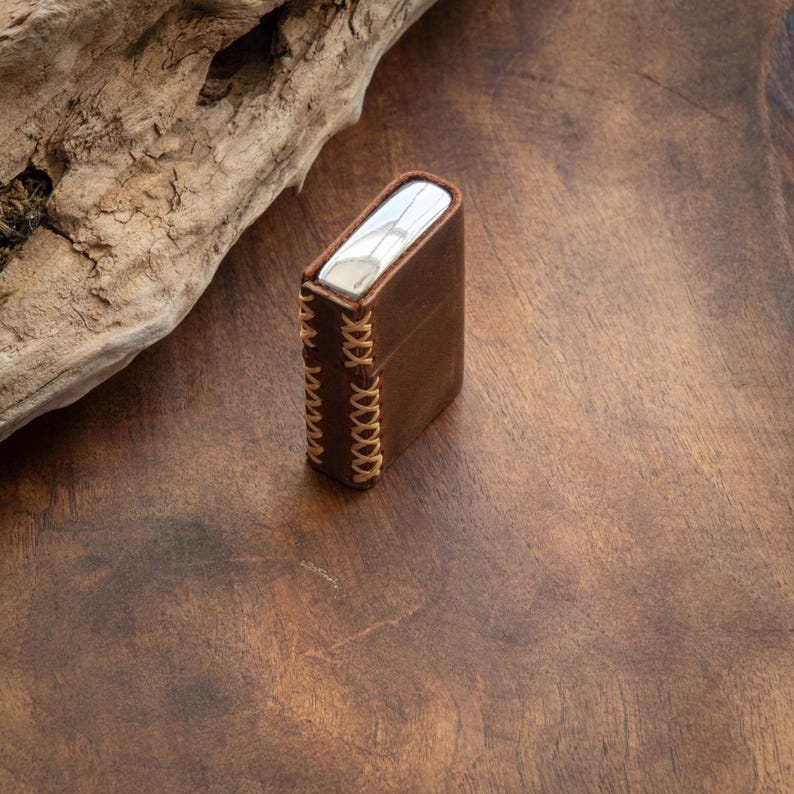May include: A brown leather case with a silver metal insert and decorative stitching. The case is sitting on a wooden surface.