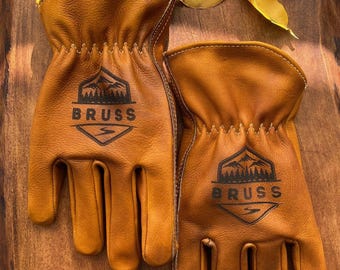 May include: A pair of tan leather work gloves with the word "BRUSS" printed in black, along with a mountain and tree logo. The gloves are stitched with a light tan thread and are resting on a wooden surface.