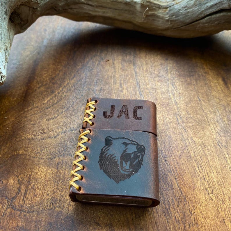 May include: A brown leather lighter case with a bear design and the name "JAC" engraved on the top. The case has a stitched edge.
