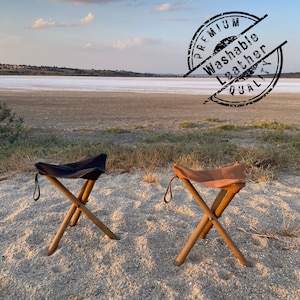 May include: Two folding stools with wooden legs and leather seats, one in black and one in tan. Each stool features a small leather loop on the side. A circular stamp on the image reads: PREMIUM Washable Leather QUALITY.