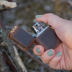 May include: A brown leather lighter case with stitching detail. The case is open, revealing a silver lighter inside.
