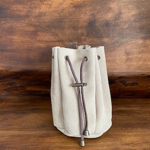 Può includere: Un sacchetto con coulisse in camoscio beige con cordoncino marrone scuro e dettagli in metallo. Il sacchetto è raccolto in alto e ha un fondo arrotondato. Su sfondo in legno scuro.