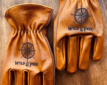 May include: A pair of tan leather work gloves. Each glove features a black compass rose design and the words "WILD & FREE". The gloves are shown on a wooden surface.