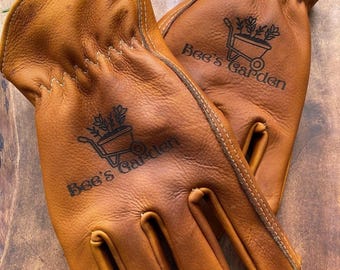 May include: A pair of tan leather gardening gloves. The gloves have a stitched design and the text "Bee's Garden" with a graphic of a wheelbarrow with plants. The gloves are made of a soft, durable leather.