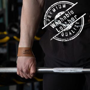 May include: A brown leather wrist wrap with a black logo of a leopard and the text "REDVARGUS". The wrap is worn on the wrist while holding a barbell. The image also includes a white stamp that says "PREMIUM Washable Leather QUALITY".