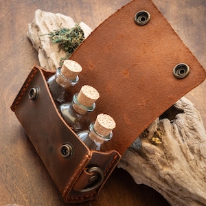 Op de afbeelding: Een bruine leren tas met drie kleine glazen flesjes met kurken stoppen. De tas is open en de flesjes zijn zichtbaar van binnen.