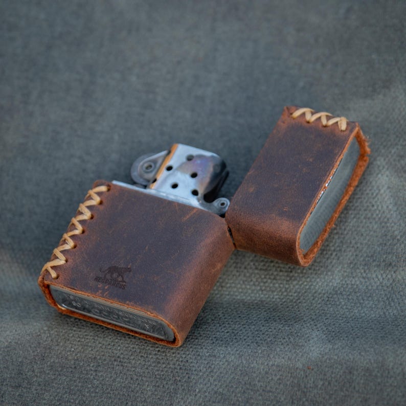 May include: A brown leather lighter case with stitching around the edges. The case is open and a silver lighter is inside. The case has the word "Adventure" embossed on it.