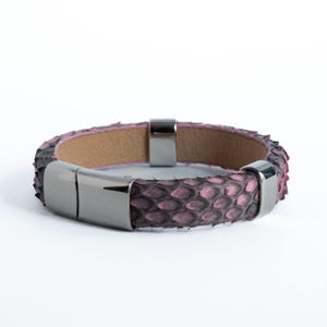 May include: A pink and black snakeskin bracelet with a magnetic clasp. The bracelet features a light brown interior and silver-colored metal clasps. The snakeskin pattern is a mix of pink and black scales.