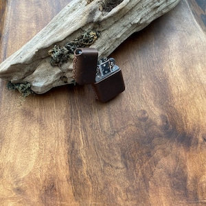 May include: A brown leather lighter case with a lighter inside, open to reveal the metal lighter. The case is stitched with a lighter brown thread. The lighter and case are on a wooden surface with a piece of driftwood.